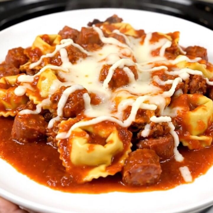 A top-down view of a slow cooker filled with browned Italian sausage, cheese tortellini, and rich tomato sauce.