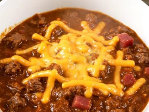 A steaming, glossy bowl of bacon cheeseburger chili topped with shredded cheddar cheese.