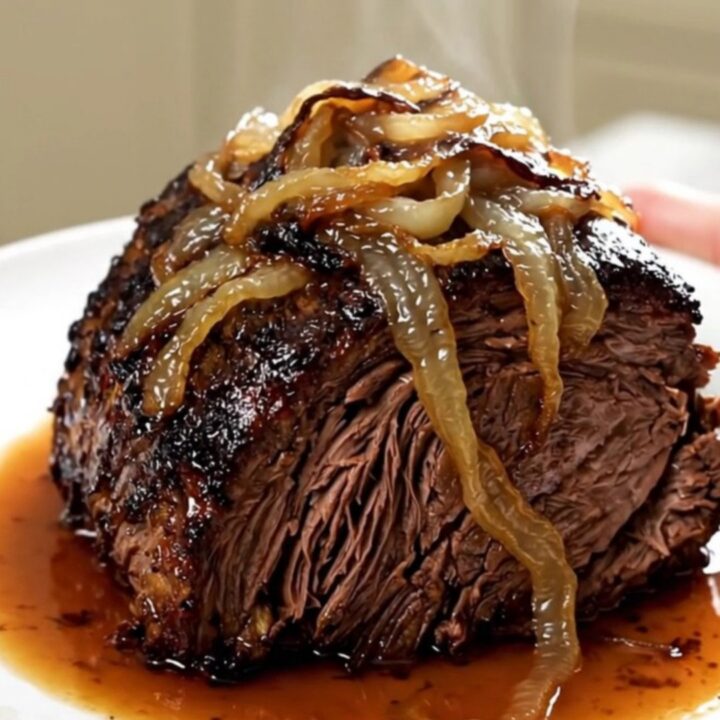 A tender, slow-cooked beef chuck roast topped with caramelized sweet onions and crispy bacon on a white plate.