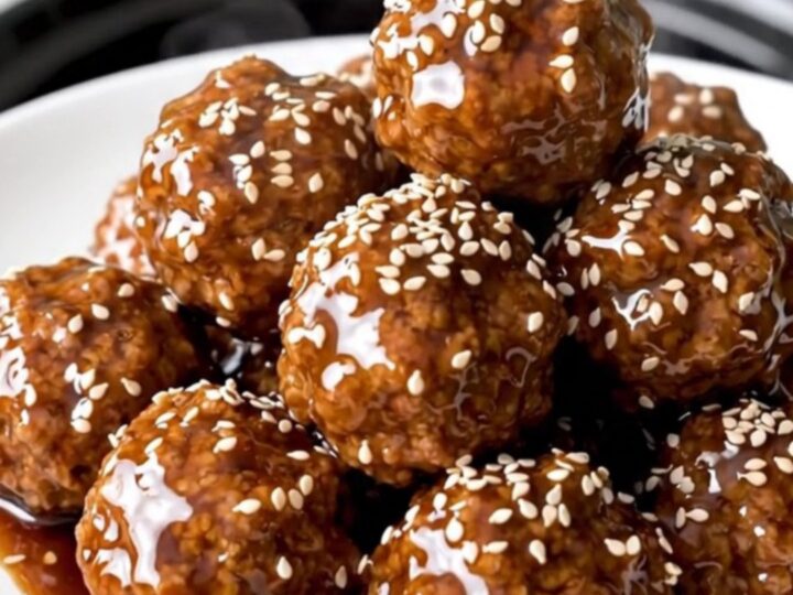 A black slow cooker filled with glazed beef meatballs and pineapple chunks on a white speckled countertop.