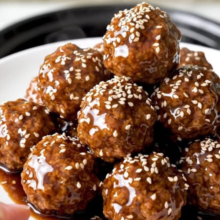 A black slow cooker filled with glazed beef meatballs and pineapple chunks on a white speckled countertop.