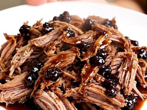 A slow cooker filled with tender pulled pork glazed in a rich blackberry sauce with visible pieces of jalapeno.