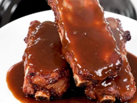 A plate of tender, glossy barbecue ribs glistening with sauce, served against a blurred kitchen background.