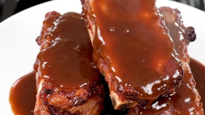 A plate of tender, glossy barbecue ribs glistening with sauce, served against a blurred kitchen background.