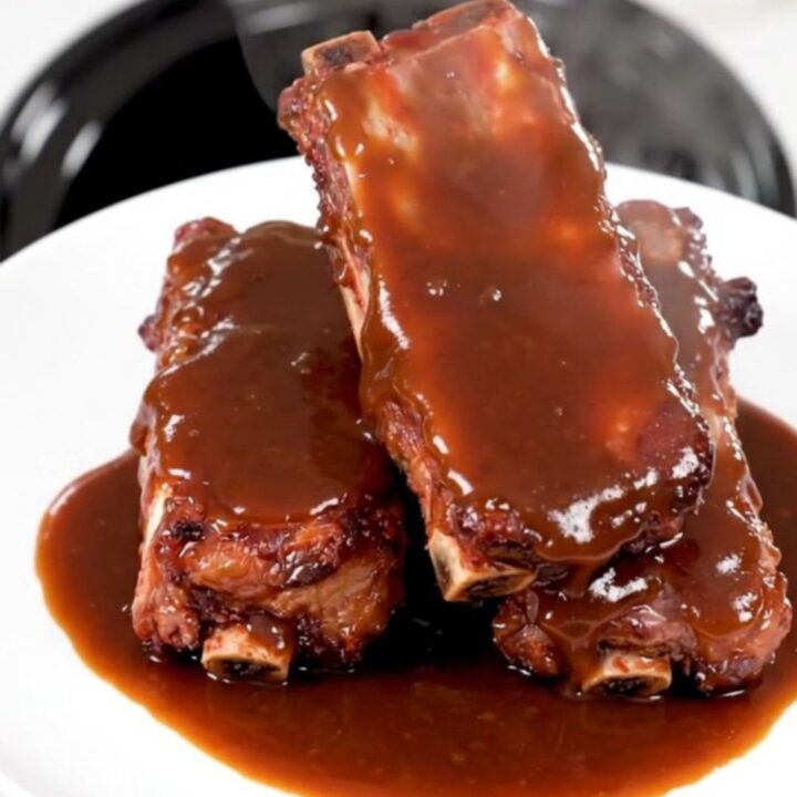 A plate of tender, glossy barbecue ribs glistening with sauce, served against a blurred kitchen background.