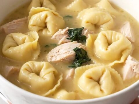 A steaming bowl of creamy chicken tortellini soup with visible pieces of chicken and fresh spinach.
