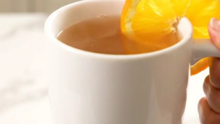 A white ceramic mug filled with steaming hot orange cinnamon wassail, garnished with an orange slice.