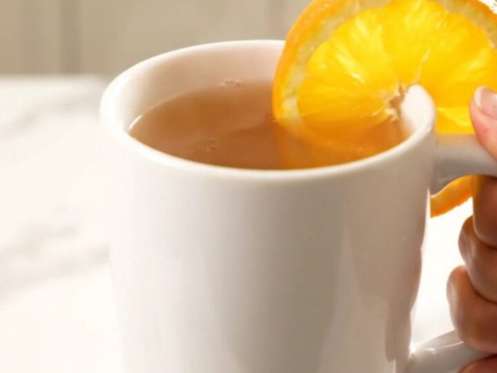 A white ceramic mug filled with steaming hot orange cinnamon wassail, garnished with an orange slice.