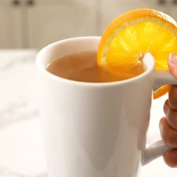 A white ceramic mug filled with steaming hot orange cinnamon wassail, garnished with an orange slice.