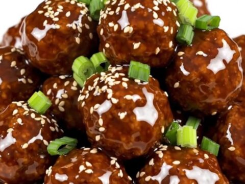 A plate of glossy, steaming General Tso meatballs served on a white round plate with a soft blurred kitchen background.