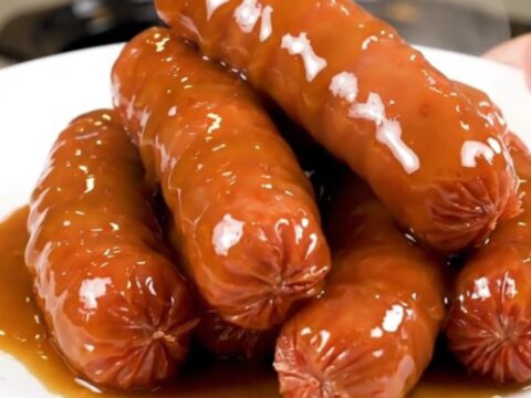 A slow cooker filled with bratwurst links, sliced onions, and a rich beer and mustard glaze on a speckled countertop.