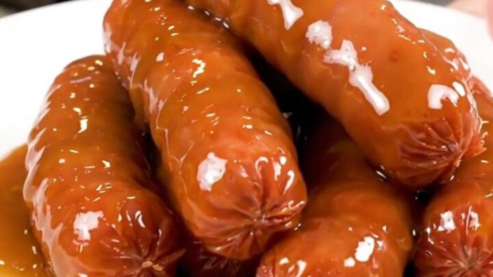 A slow cooker filled with bratwurst links, sliced onions, and a rich beer and mustard glaze on a speckled countertop.