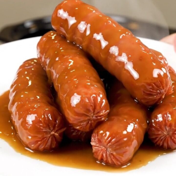 A slow cooker filled with bratwurst links, sliced onions, and a rich beer and mustard glaze on a speckled countertop.