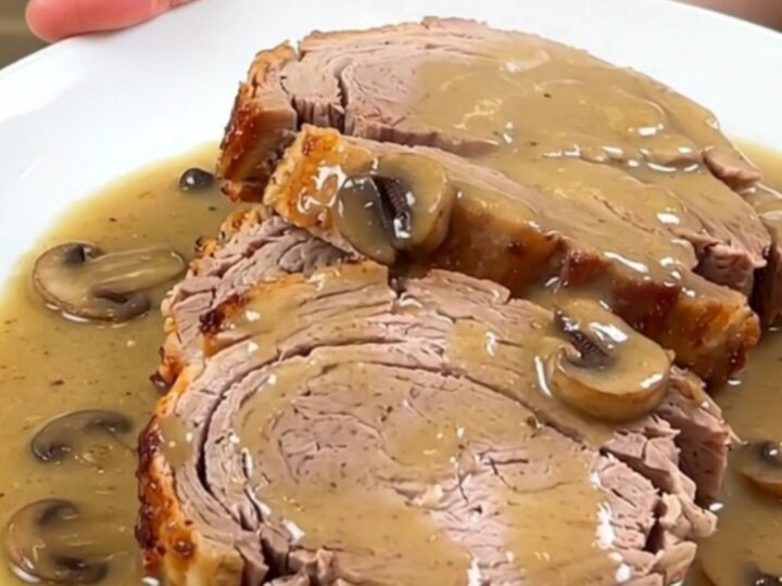 A close-up of a tender sliced pork loin smothered in a rich, glossy creamy mushroom gravy on a white plate.
