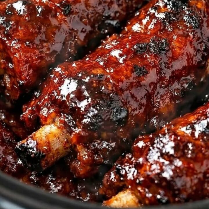 A close-up shot of tender, sticky honey garlic BBQ beef ribs glistening under warm kitchen lights inside a slow cooker.