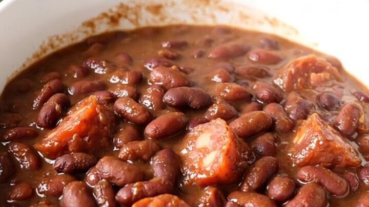 A slow cooker filled with hearty charro beans, chorizo, and onions on a white quartz countertop.