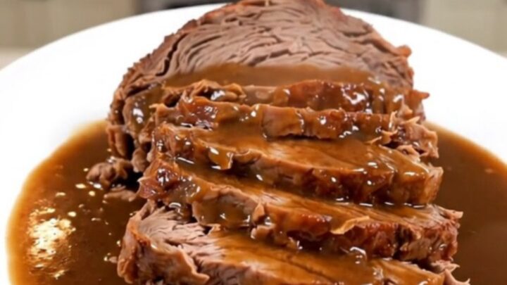 A close-up of a tender, sliced pot roast with glossy horseradish gravy on a white plate.