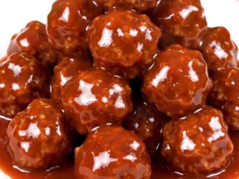 A black slow cooker filled with glossy, dark-glazed meatballs on a white speckled countertop.