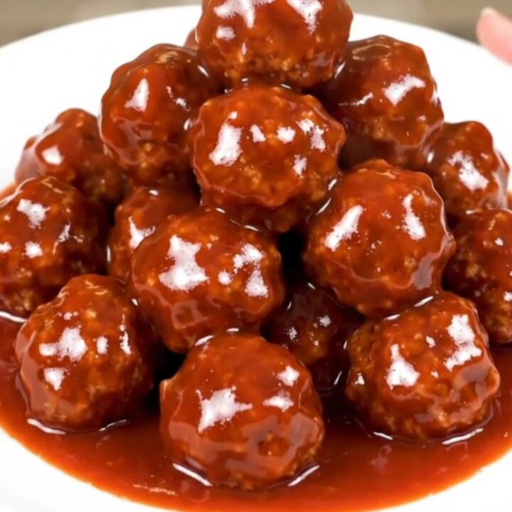 A black slow cooker filled with glossy, dark-glazed meatballs on a white speckled countertop.