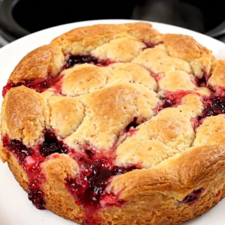 A top-down view of a black oval slow cooker filled with a bubbling apple and berry cobbler, topped with melted butter.