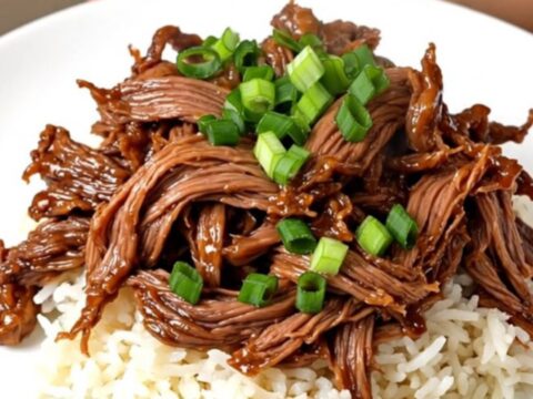A close-up view of tender, glossy teriyaki shredded beef served over white jasmine rice with scallion garnish.