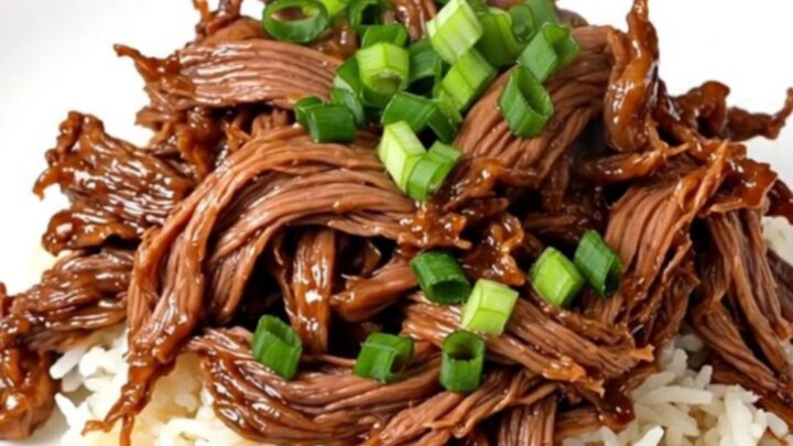 A close-up view of tender, glossy teriyaki shredded beef served over white jasmine rice with scallion garnish.