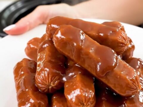 A top-down view into a black slow cooker filled with glazed cocktail sausages in a thick