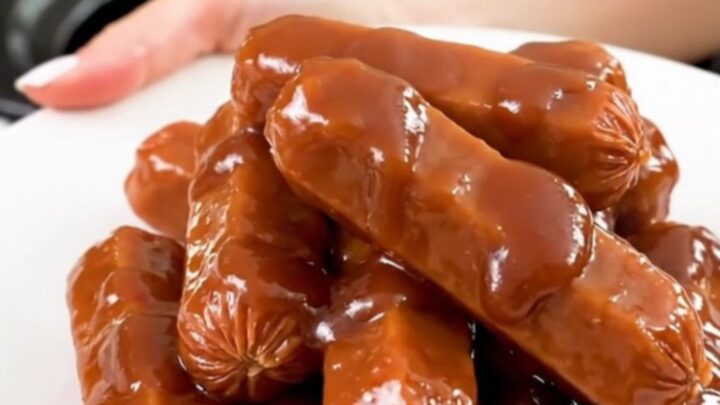 A top-down view into a black slow cooker filled with glazed cocktail sausages in a thick