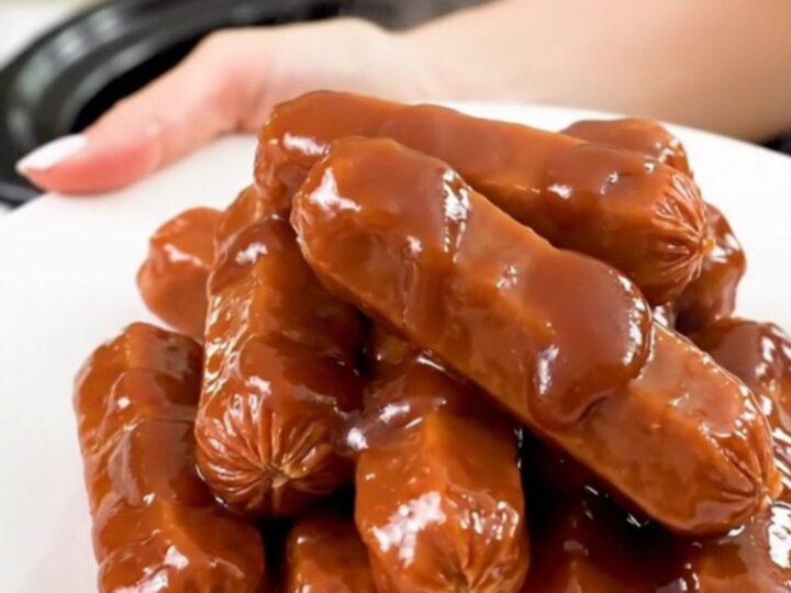 A top-down view into a black slow cooker filled with glazed cocktail sausages in a thick