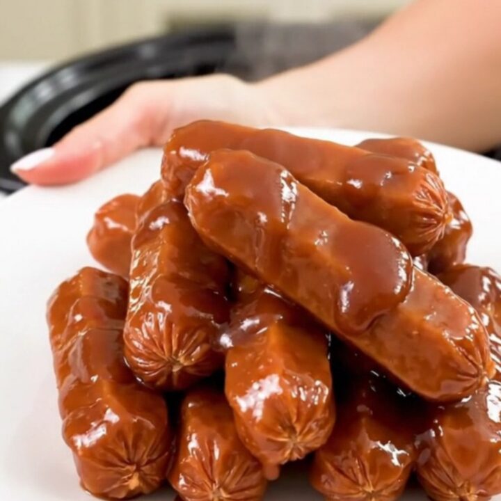 A top-down view into a black slow cooker filled with glazed cocktail sausages in a thick
