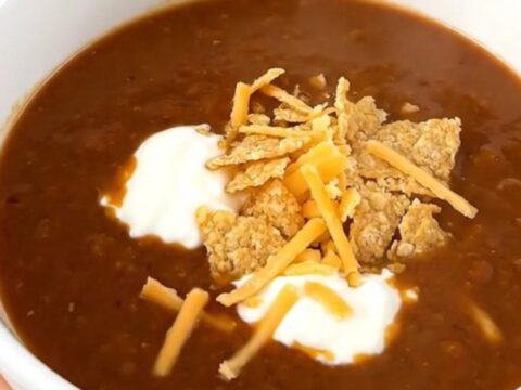 A steaming bowl of ground beef enchilada soup topped with sour cream, shredded cheese, and tortilla chips.
