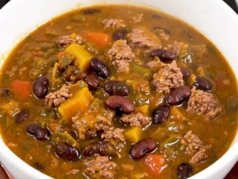 A white bowl filled with glossy, dark red beef and black bean chili, held in front of blurred kitchen cabinets.