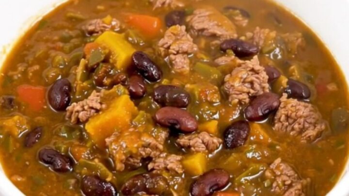 A white bowl filled with glossy, dark red beef and black bean chili, held in front of blurred kitchen cabinets.