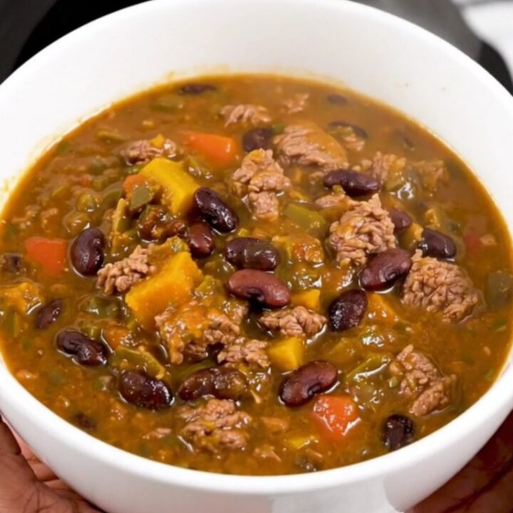 A white bowl filled with glossy, dark red beef and black bean chili, held in front of blurred kitchen cabinets.
