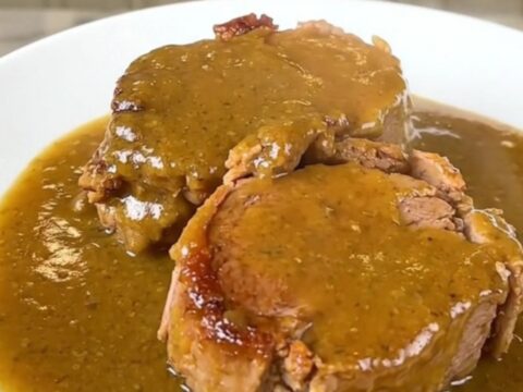 A close-up of a tender, slow-cooked pork roast smothered in a rich golden mushroom gravy on a white plate.