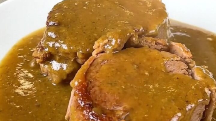 A close-up of a tender, slow-cooked pork roast smothered in a rich golden mushroom gravy on a white plate.