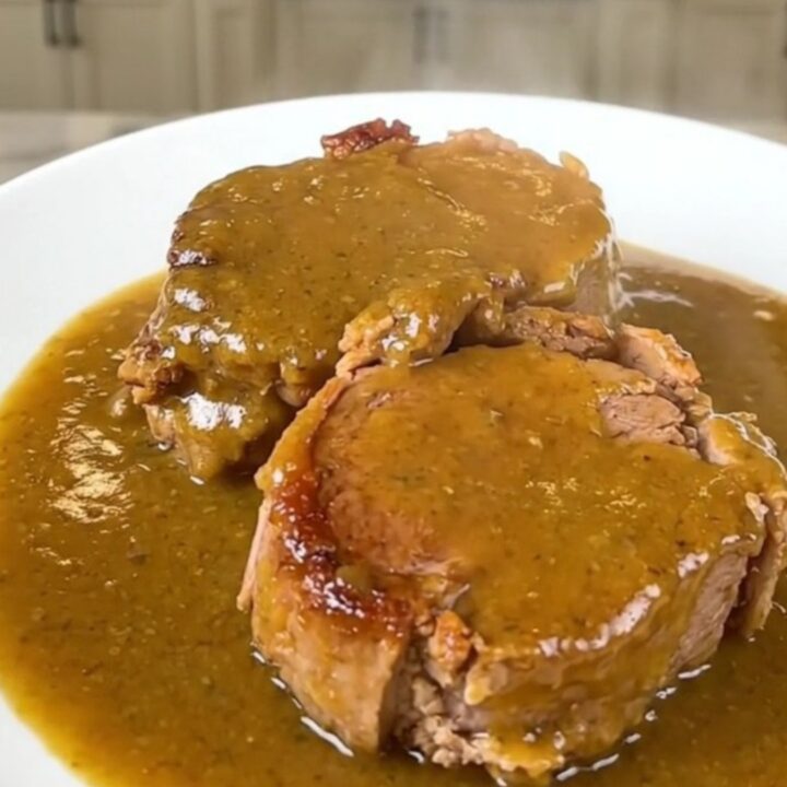 A close-up of a tender, slow-cooked pork roast smothered in a rich golden mushroom gravy on a white plate.