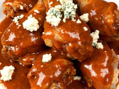 A top-down view of a black slow cooker filled with raw chicken drumsticks, celery, and buffalo sauce.
