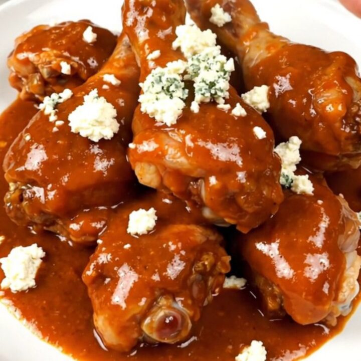 A top-down view of a black slow cooker filled with raw chicken drumsticks, celery, and buffalo sauce.