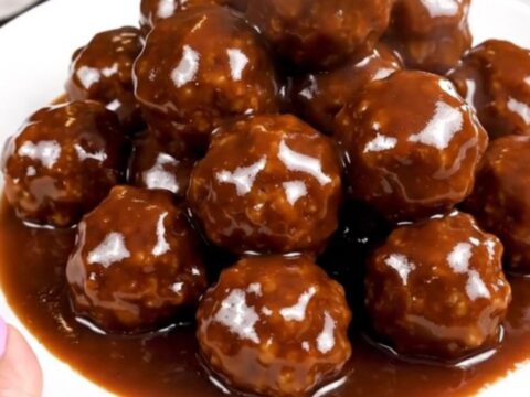 A close-up of glossy, dark-glazed meatballs in a slow cooker on a white speckled countertop.