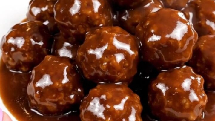 A close-up of glossy, dark-glazed meatballs in a slow cooker on a white speckled countertop.