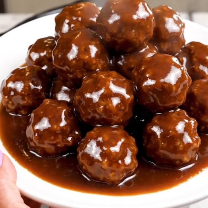 A close-up of glossy, dark-glazed meatballs in a slow cooker on a white speckled countertop.