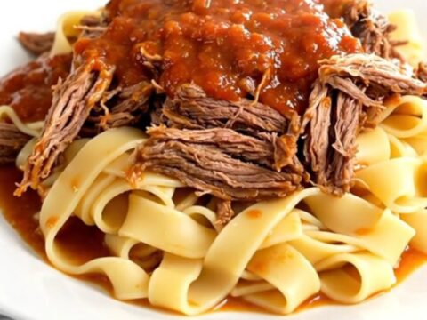 A steaming, tender beef pot roast served over pappardelle pasta on a white plate.