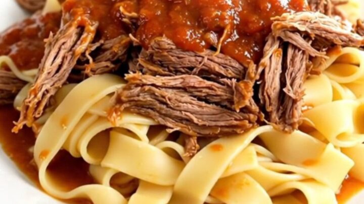A steaming, tender beef pot roast served over pappardelle pasta on a white plate.