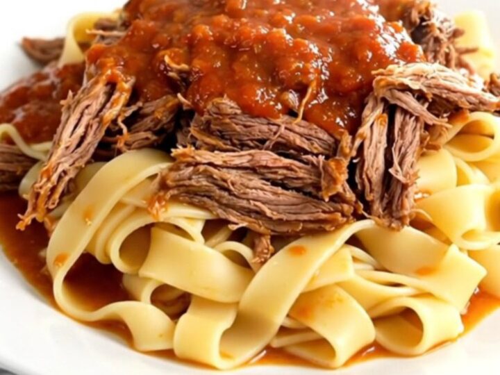 A steaming, tender beef pot roast served over pappardelle pasta on a white plate.