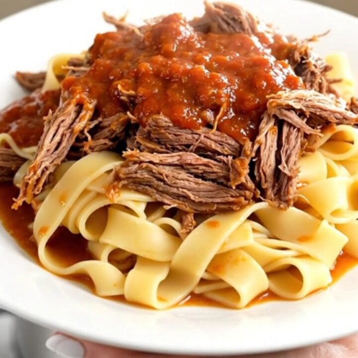 A steaming, tender beef pot roast served over pappardelle pasta on a white plate.