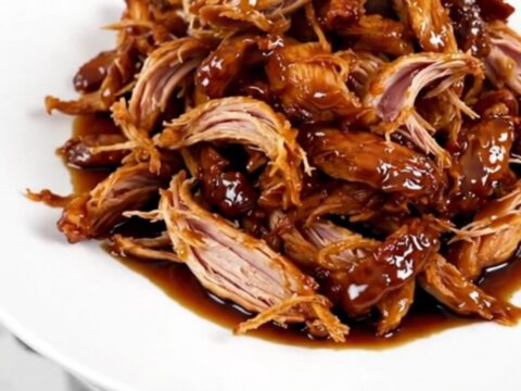 A close-up of glossy, shredded teriyaki chicken served on a white plate with a dark background.
