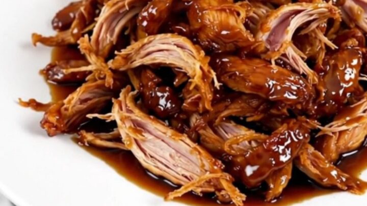A close-up of glossy, shredded teriyaki chicken served on a white plate with a dark background.