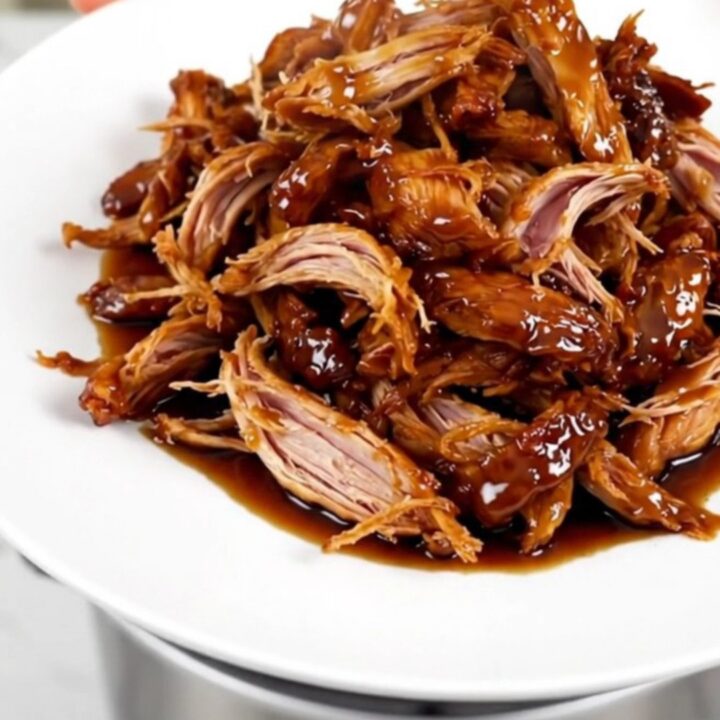 A close-up of glossy, shredded teriyaki chicken served on a white plate with a dark background.