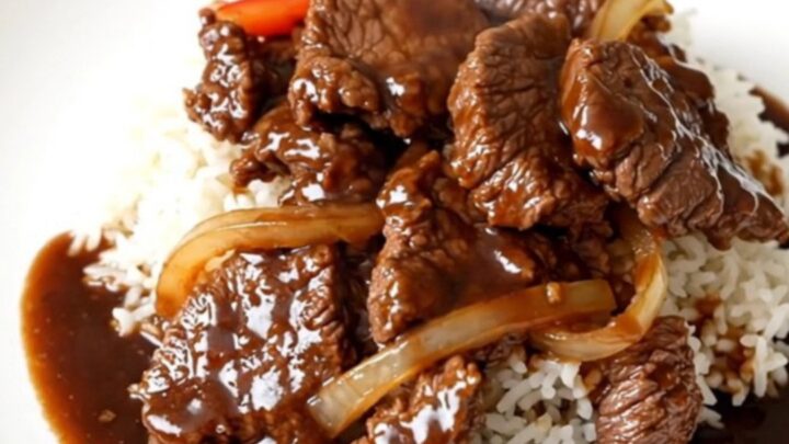 A top-down view of a slow cooker filled with tender beef strips, sliced bell peppers, and onions in a rich, glossy sauce.